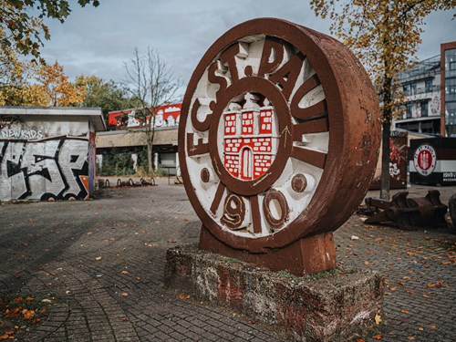 Ein großes Logo vom FC St. Pauli steht draußen. Dahinter sind einige Gebäude.