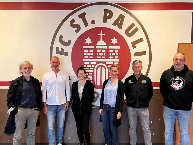 Vier Männer und zwei Frauen stehen vor einer Wand. Auf der Wand ist das Logo vom FC St. Pauli.