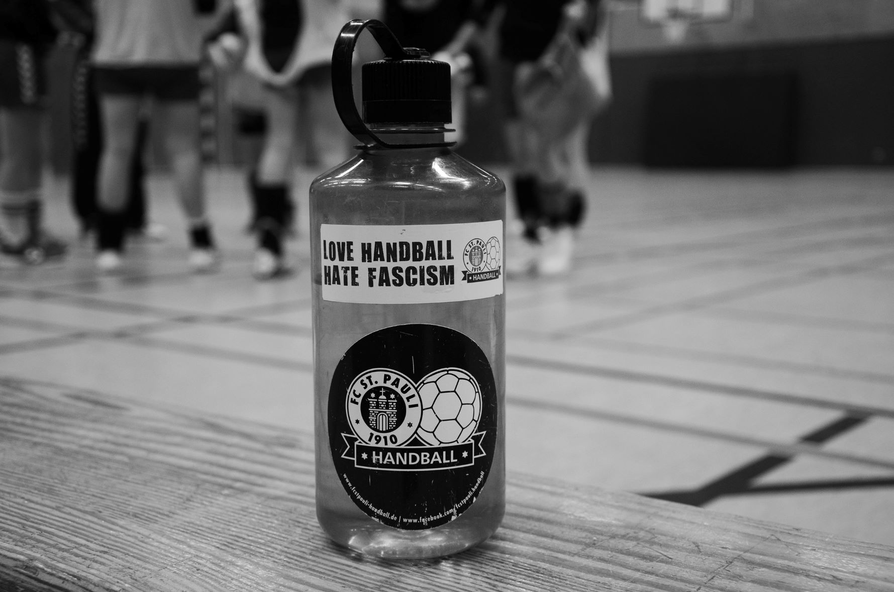Eine Trinkflasche steht in einer Sporthalle. Darauf steht: Love Handball, hate Fascism.